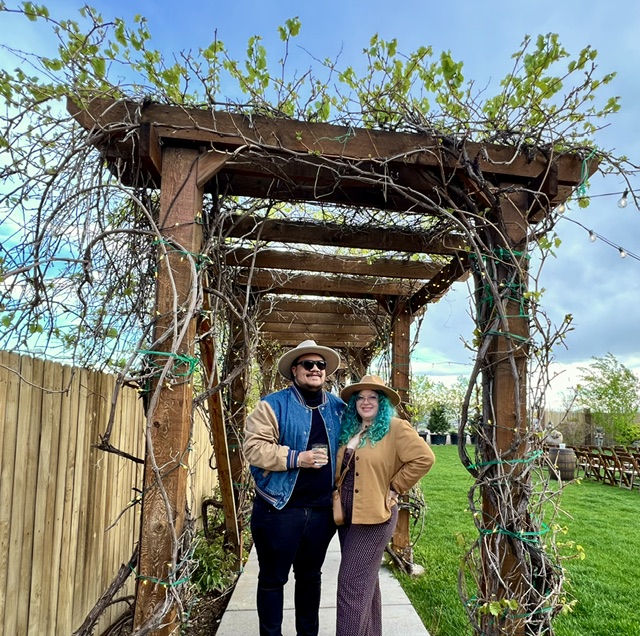 Two people stand beneath a wooden garden arbor covered with vines and string lights.