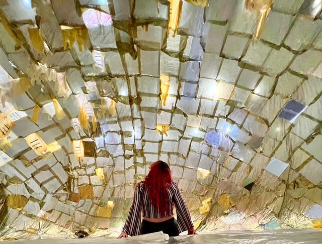 Person with long red hair stands beneath a dome covered in reflective paper pieces.