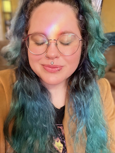 Person with teal hair and round glasses, eyes closed, wearing a tan jacket.