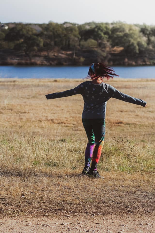 Person with outstretched arms standing in a grassy field beside a lake.