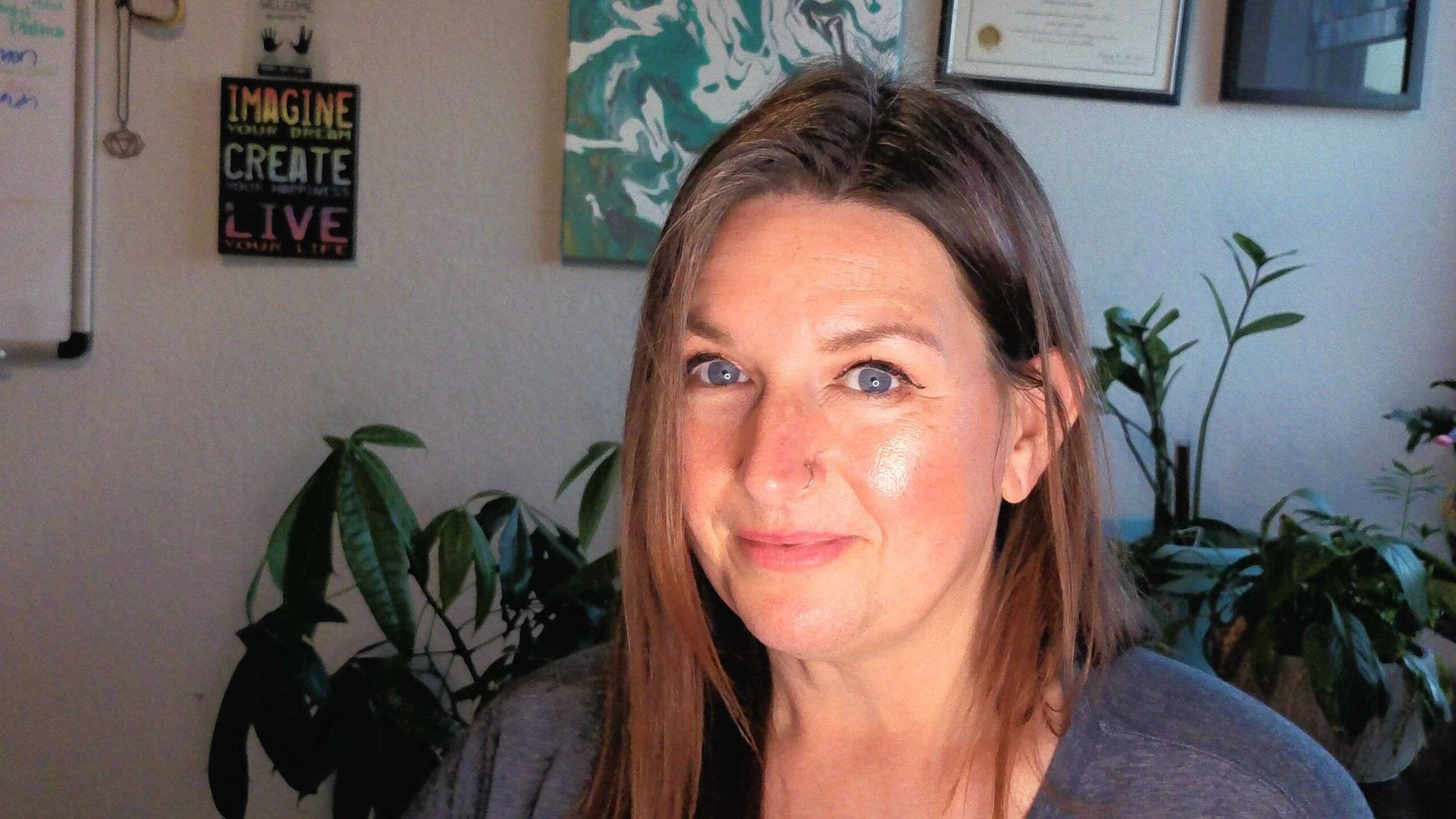 A woman with long brown hair sits in an office with plants, framed artwork, and a wall sign behind her.
