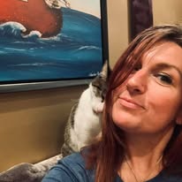 A person with reddish-brown hair poses beside a white and gray cat, with a framed ocean painting behind them.