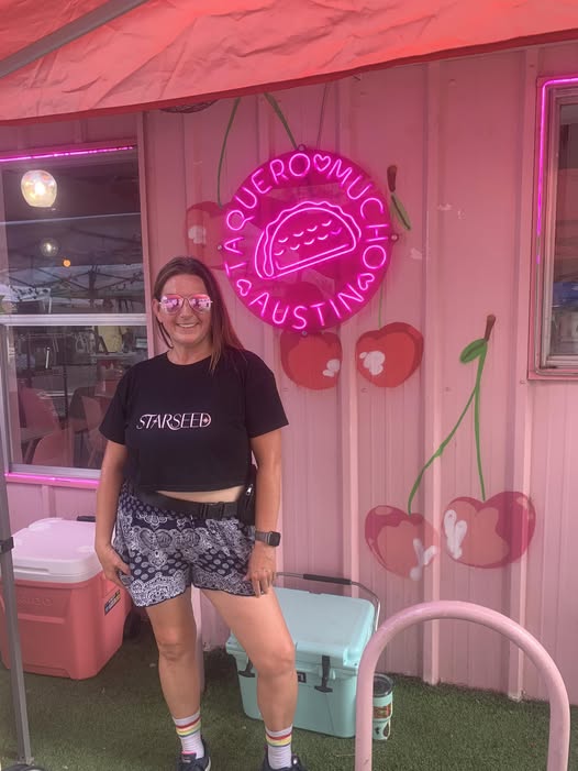 A person stands beside a neon circular sign on a pink wall, with flower decorations and colorful containers nearby.