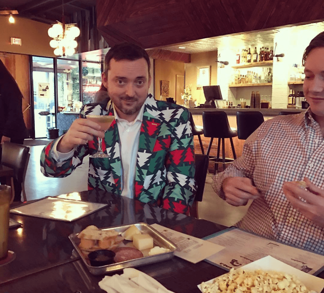 Two people sit at a restaurant table with food and drinks; one holds up a glass.