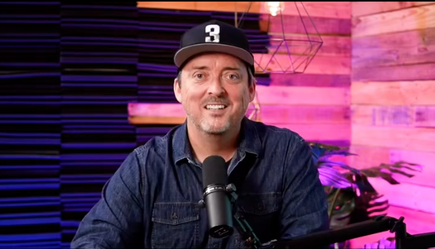 A person in a black cap sits at a microphone in a studio with dark acoustic panels and pink lighting.