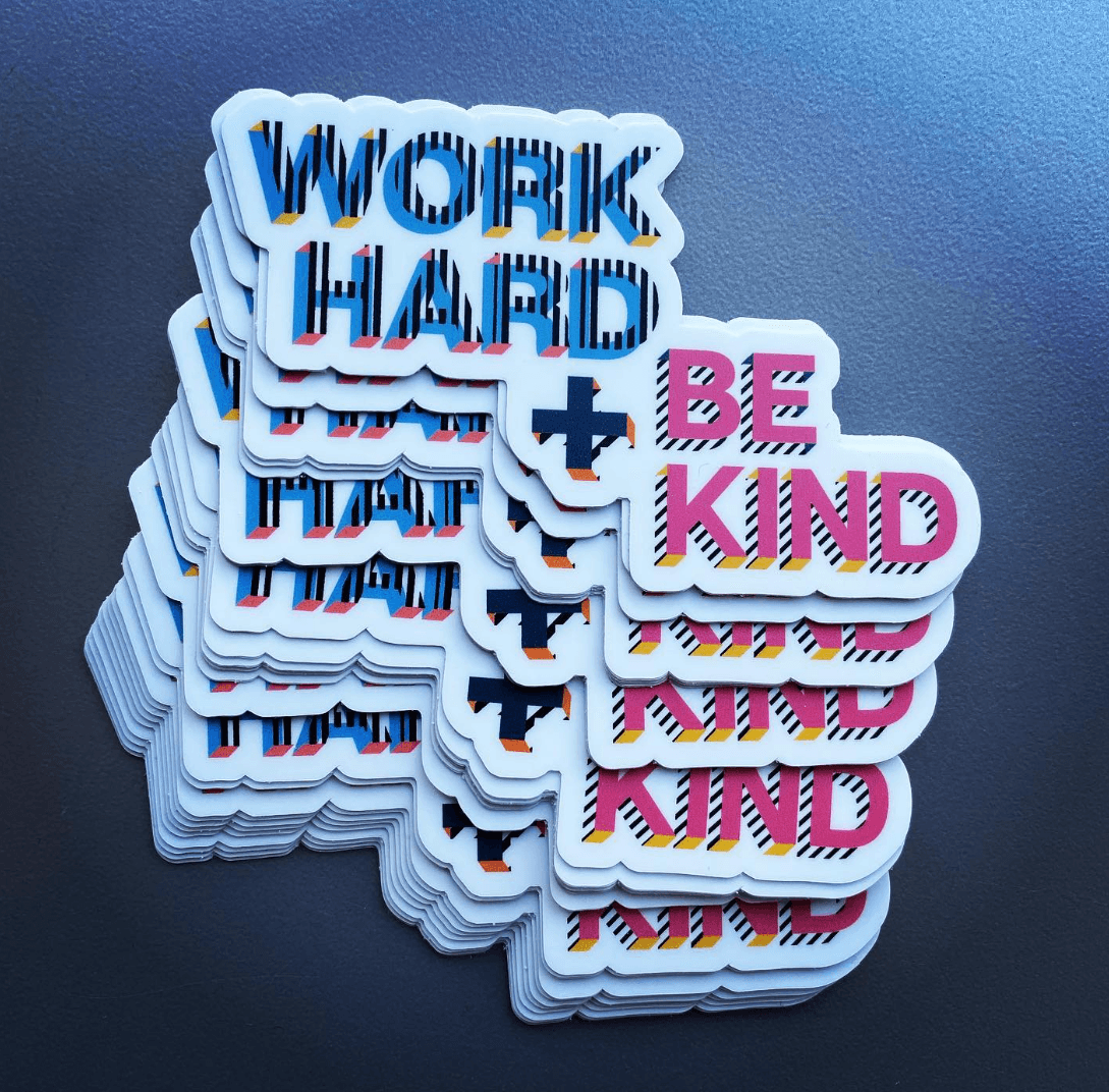 Stack of colorful stickers reading “WORK HARD” and “BE KIND” on a dark surface.