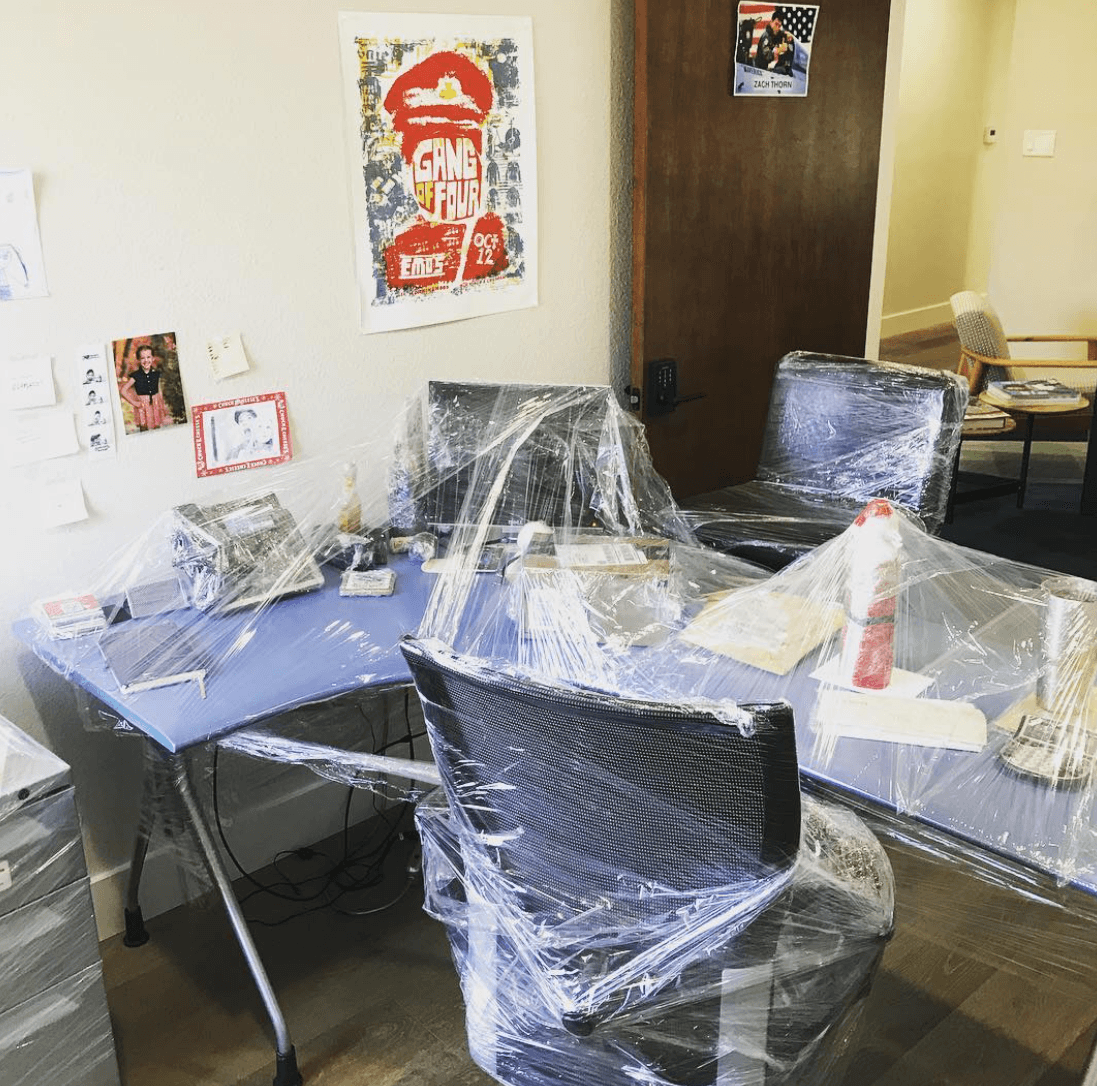 Office table and chairs wrapped in clear plastic, with papers and posters on the walls.