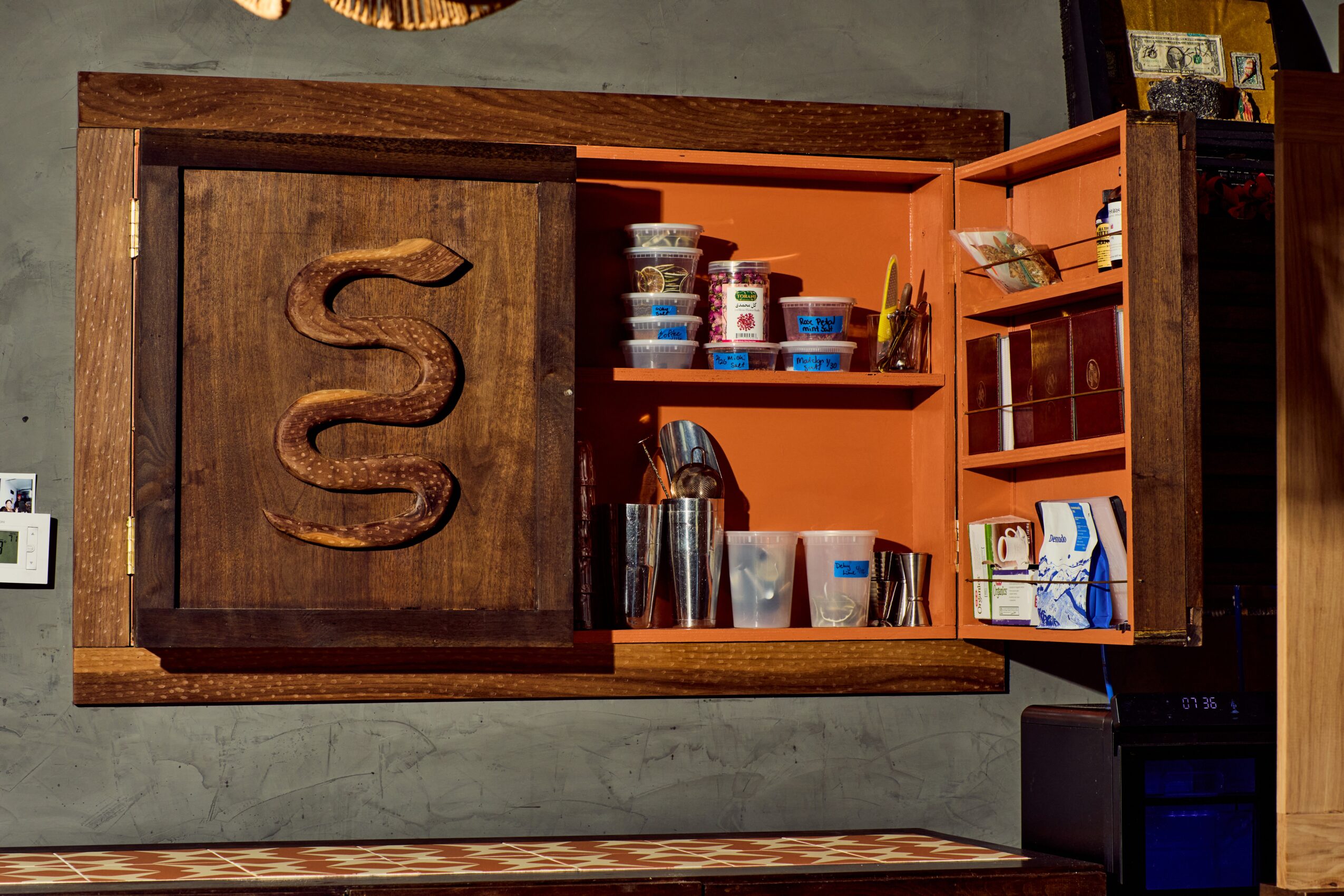 Open wooden cabinet with orange shelves, glassware, containers, and a carved snake design on the door.