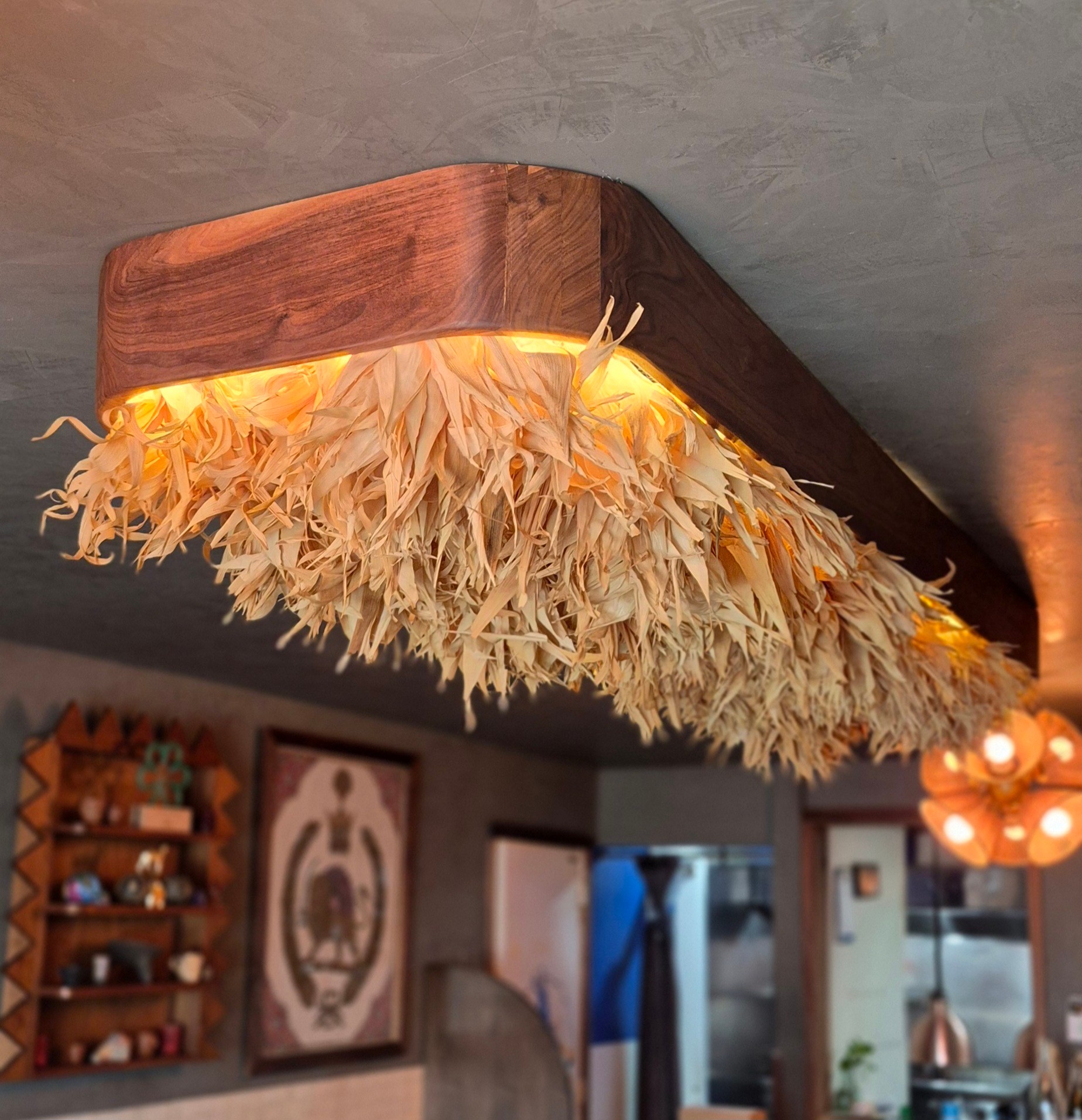 A rectangular wooden ceiling fixture glows above a restaurant interior, with hanging dried plant material beneath it.