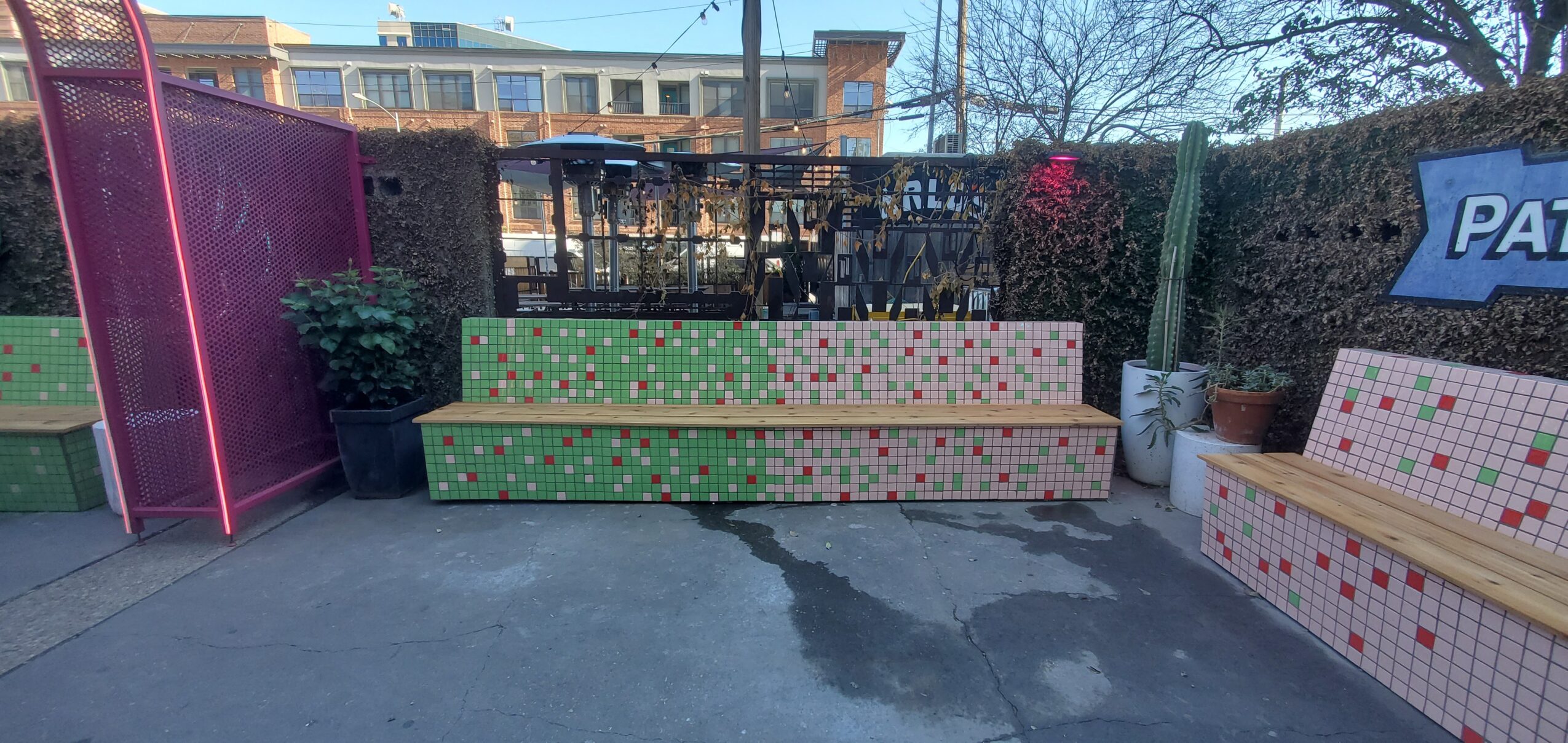 Outdoor patio with colorful tiled benches, tables, a hedge, and a building in the background.