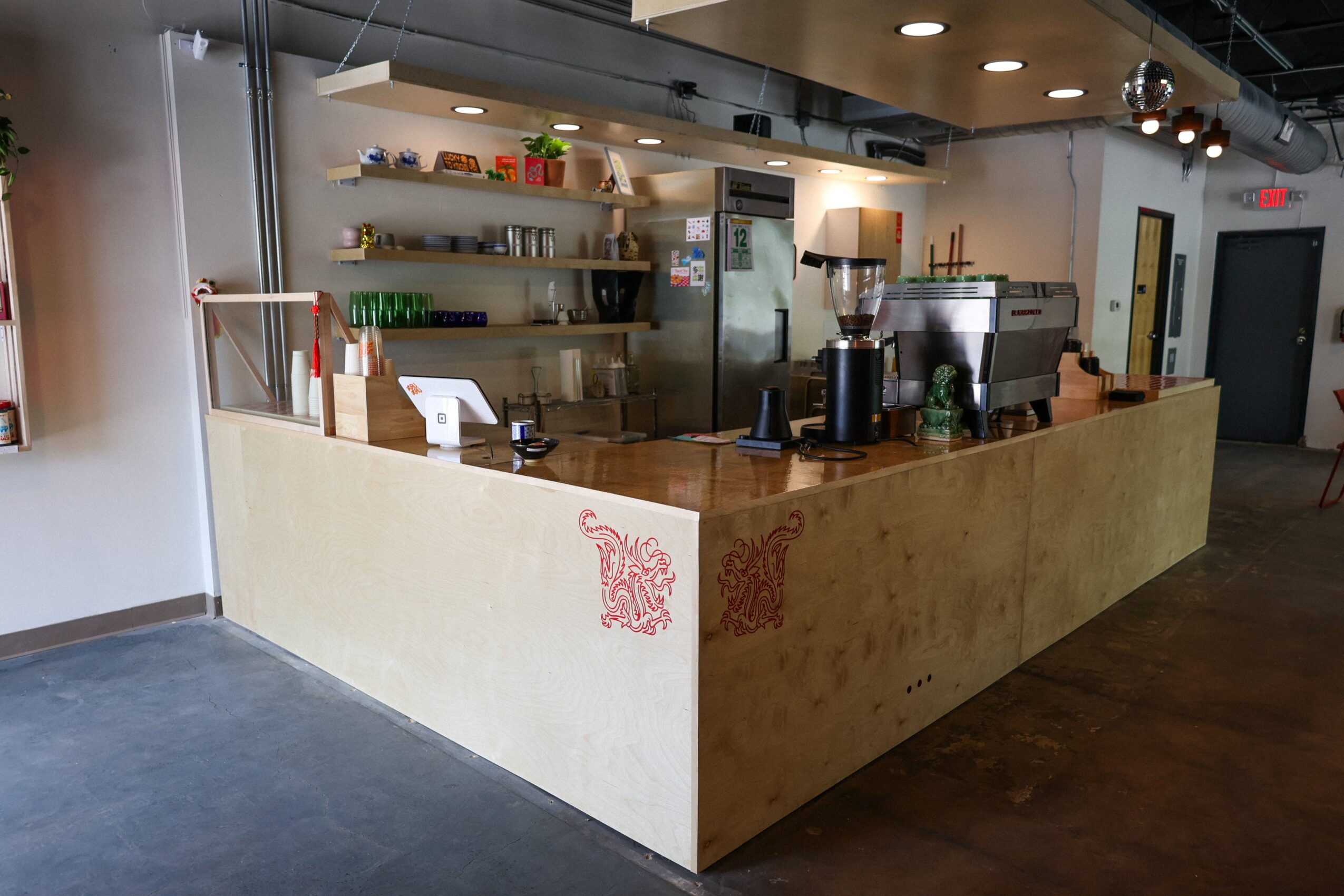 A café counter with a wooden front, espresso machine, shelves, and seating area.