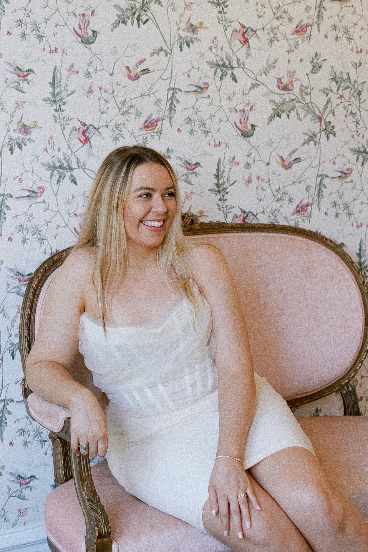 A person in a white dress sits on a pink upholstered chair against floral wallpaper.