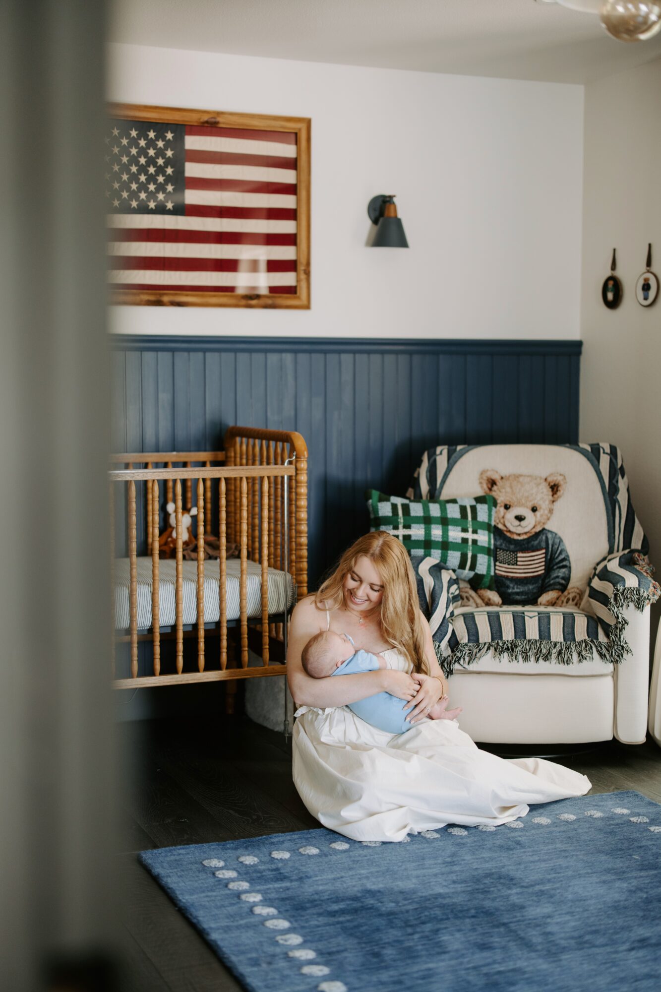 A person sits on the floor beside a crib and armchair beneath a framed American flag.