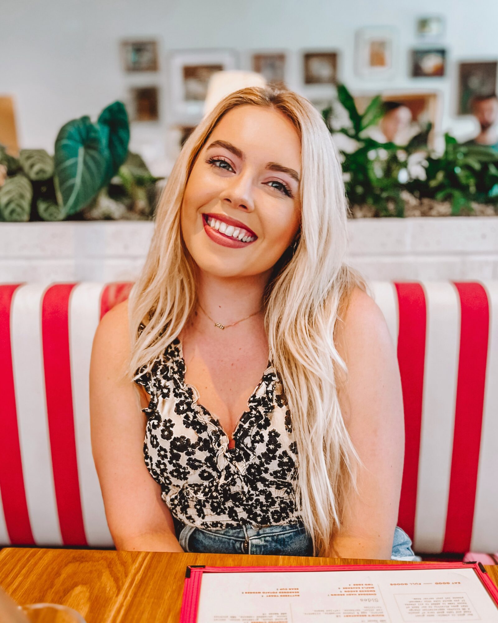 Smiling blonde person seated against a red-and-white striped booth, with plants and framed pictures behind them.