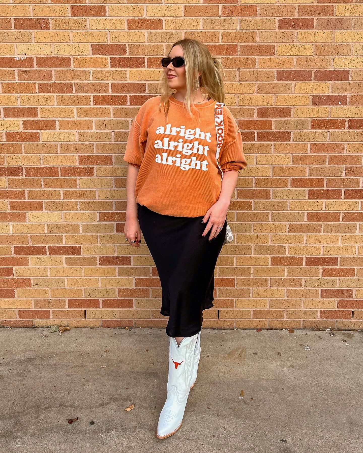 Person wearing sunglasses, an orange graphic shirt, black pants, and white boots standing against a brick wall.