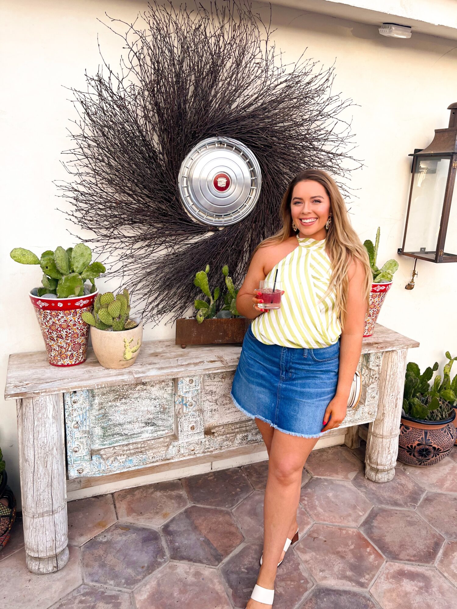A person stands beside a rustic console table and potted plants, with a large circular wall decoration behind them.