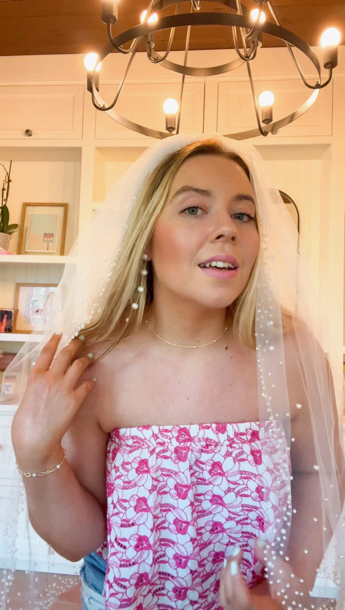 Person wearing a white veil and pink floral dress beneath a chandelier.