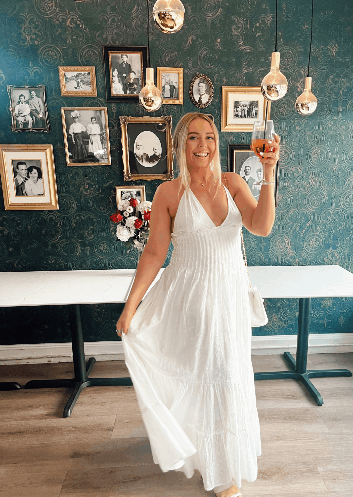 Person in a white dress holding a glass beneath hanging lights, with framed pictures on a patterned wall.