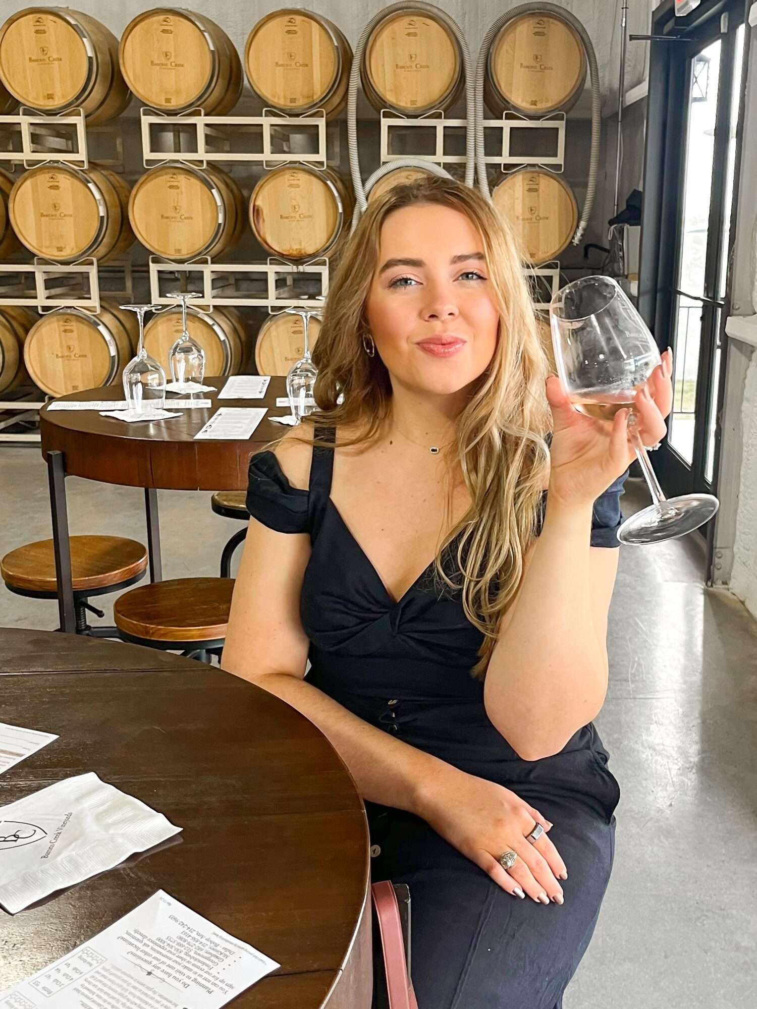 A seated person holds up a wine glass at a table, with wooden barrels lining the wall behind them.