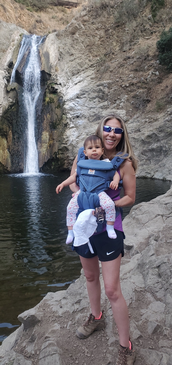 A person carries a small child beside a pool beneath a waterfall.