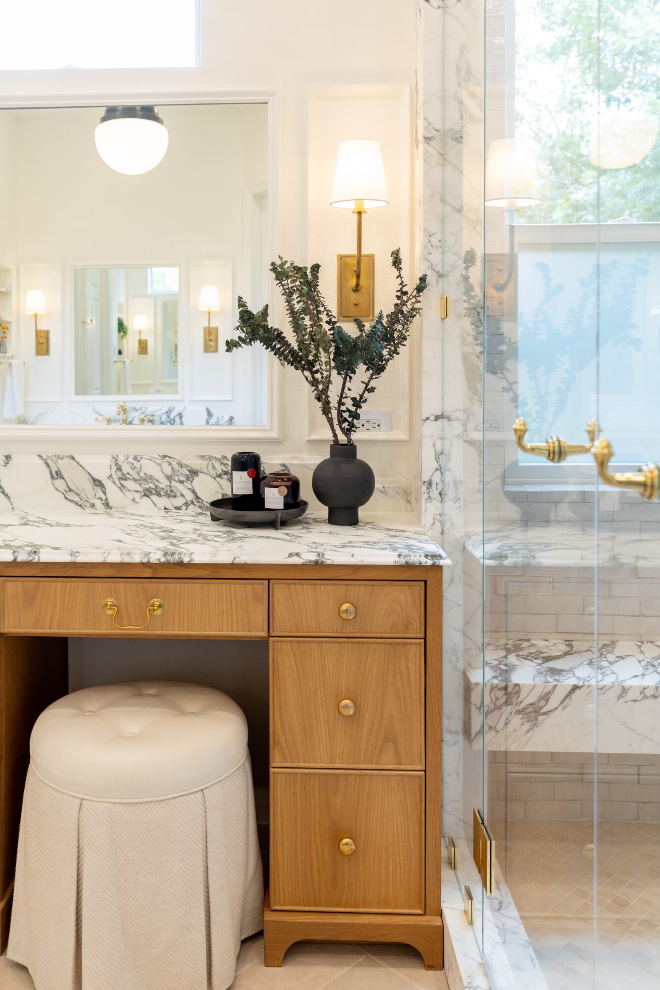 Marble bathroom vanity with a round stool, vase of branches, wall sconces, mirror, and glass shower enclosure.