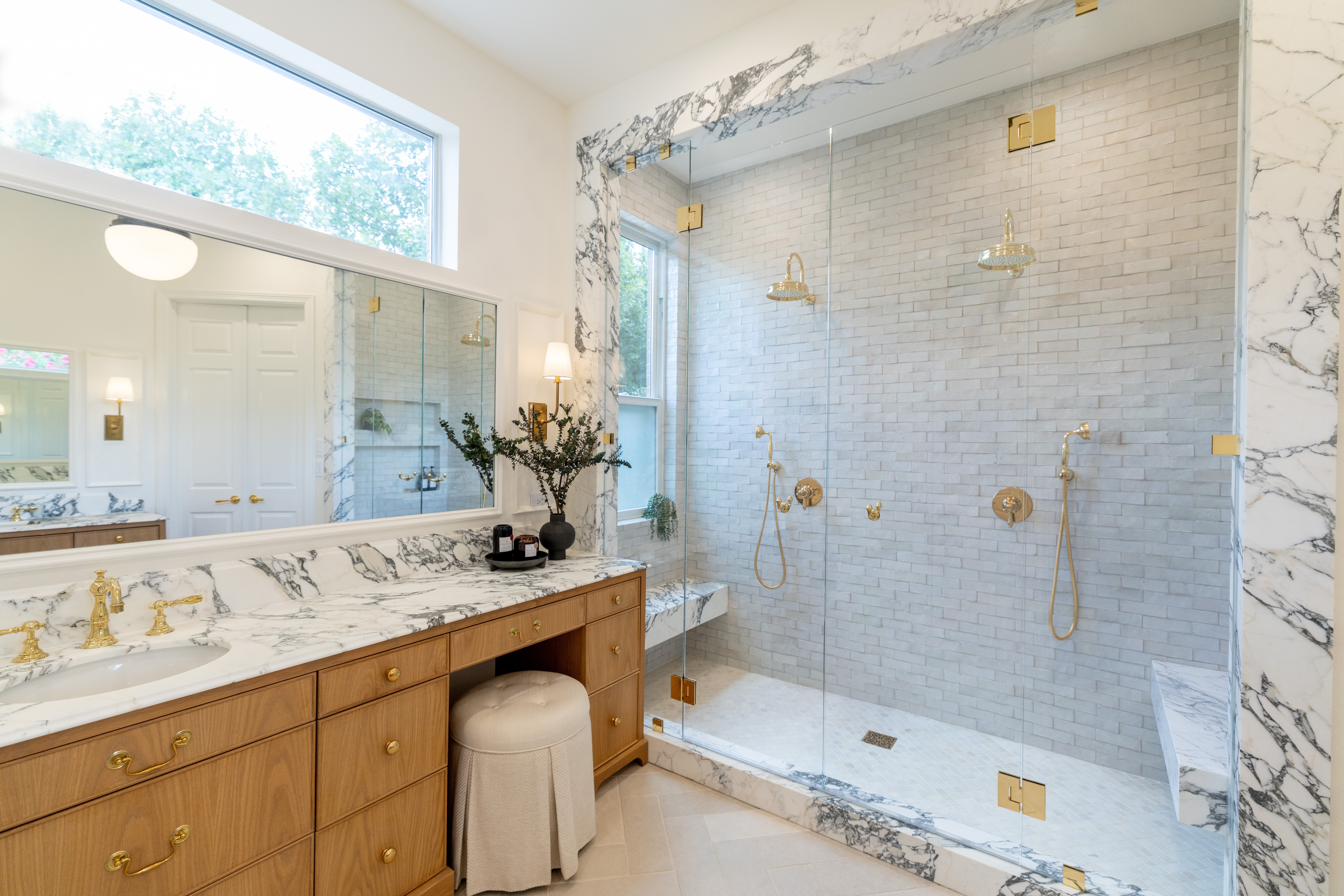 Marble bathroom with a glass-enclosed shower, wooden vanity, stool, and wide mirror beneath a high window.