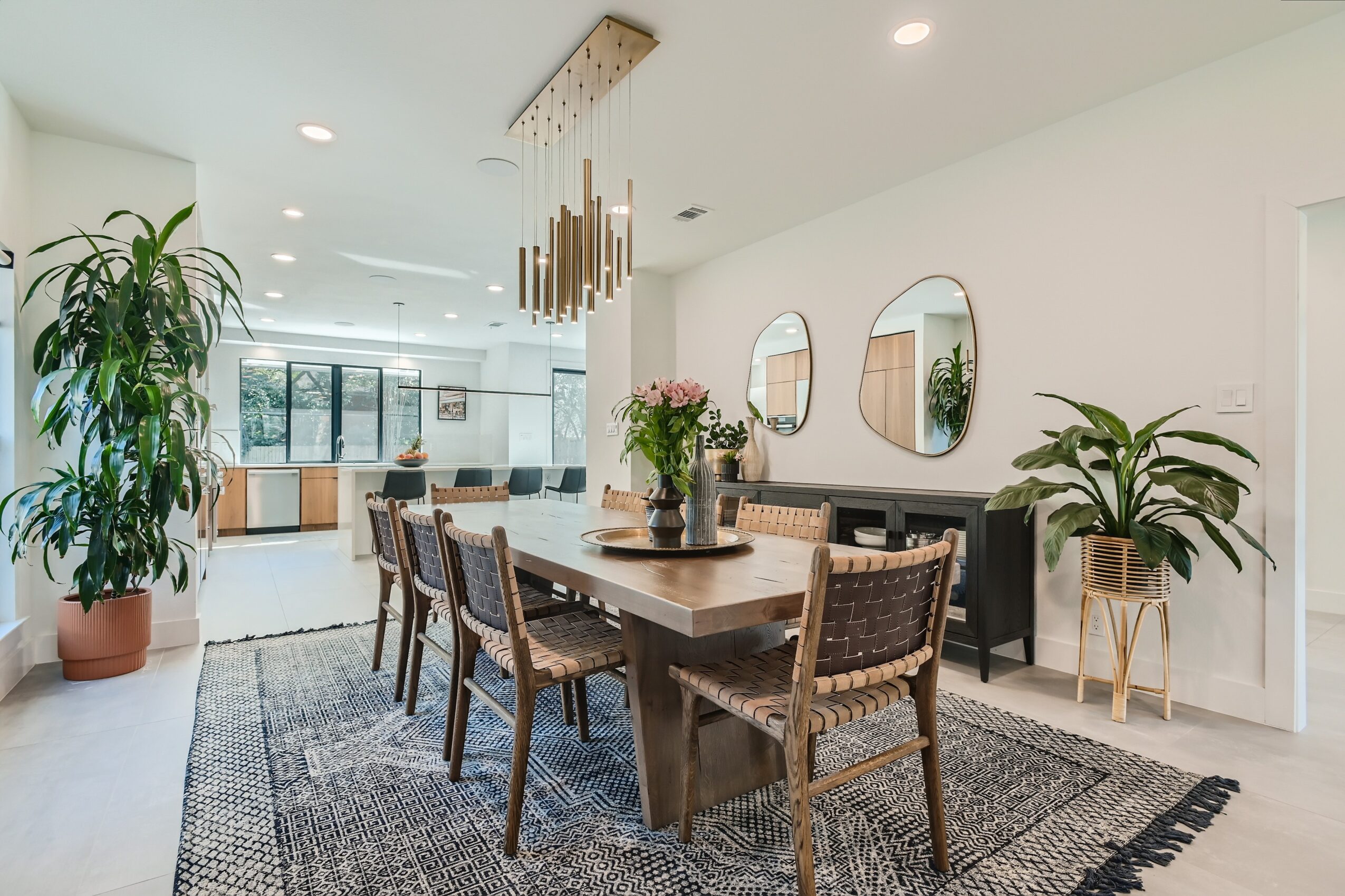 Bright dining area with a wooden table, woven chairs, pendant lights, plants, and a patterned rug.