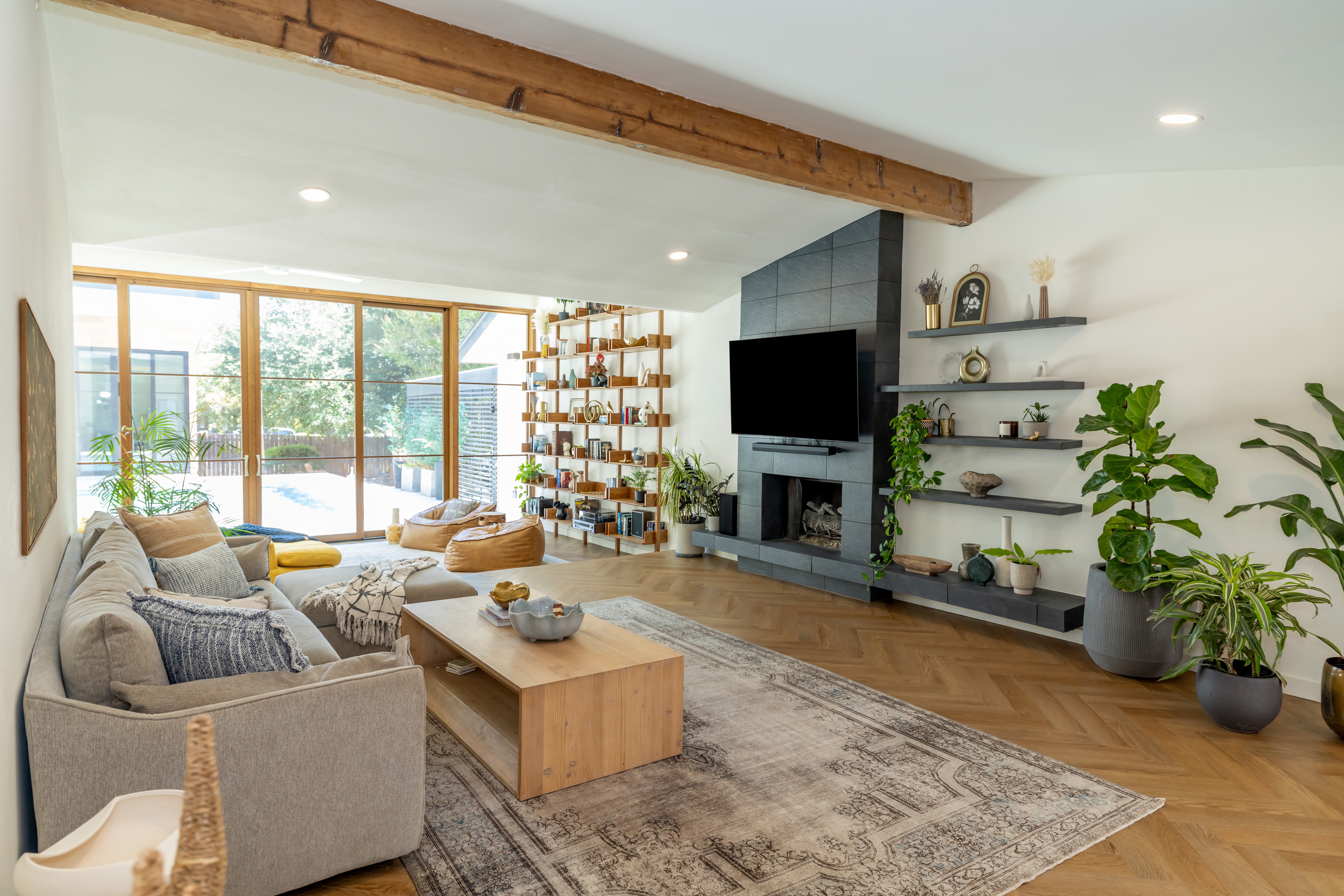Bright living room with a gray sofa, wooden coffee table, large windows, shelving, television, and potted plants.