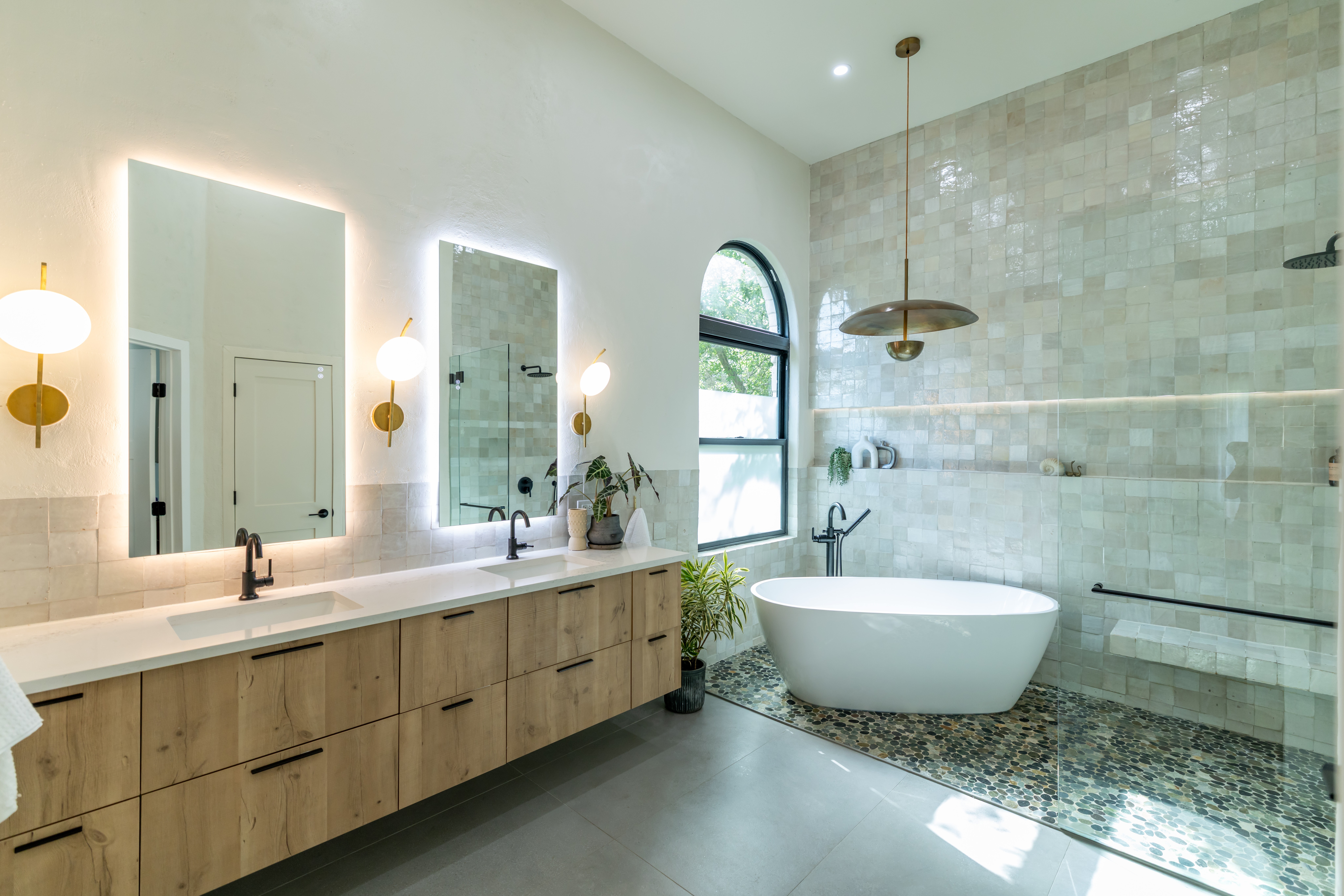 Bright bathroom with a long double vanity, mirrors, a freestanding tub, and an arched window.