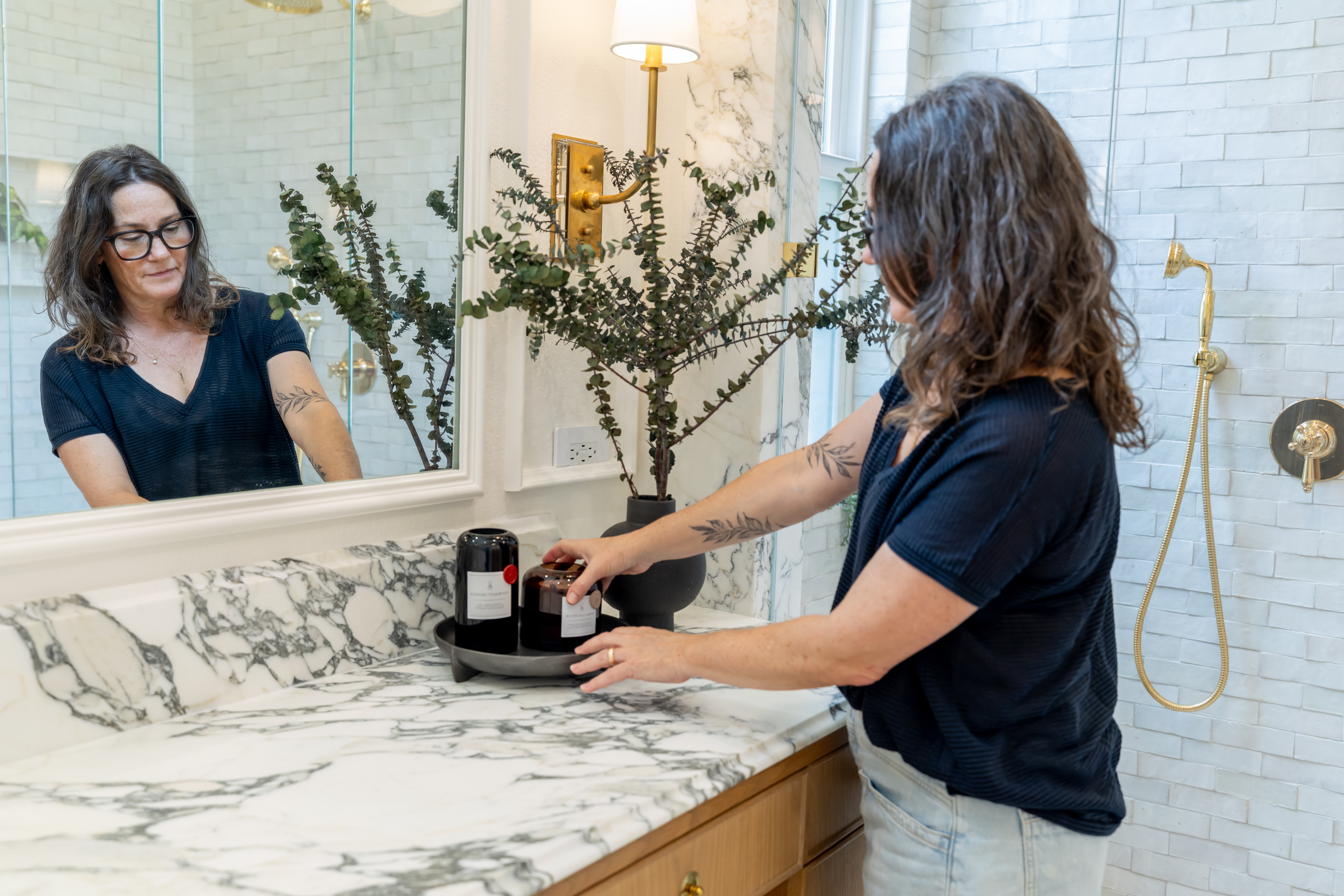 Person standing at a bathroom vanity, reflected in a mirror, reaching toward bottles on the counter.