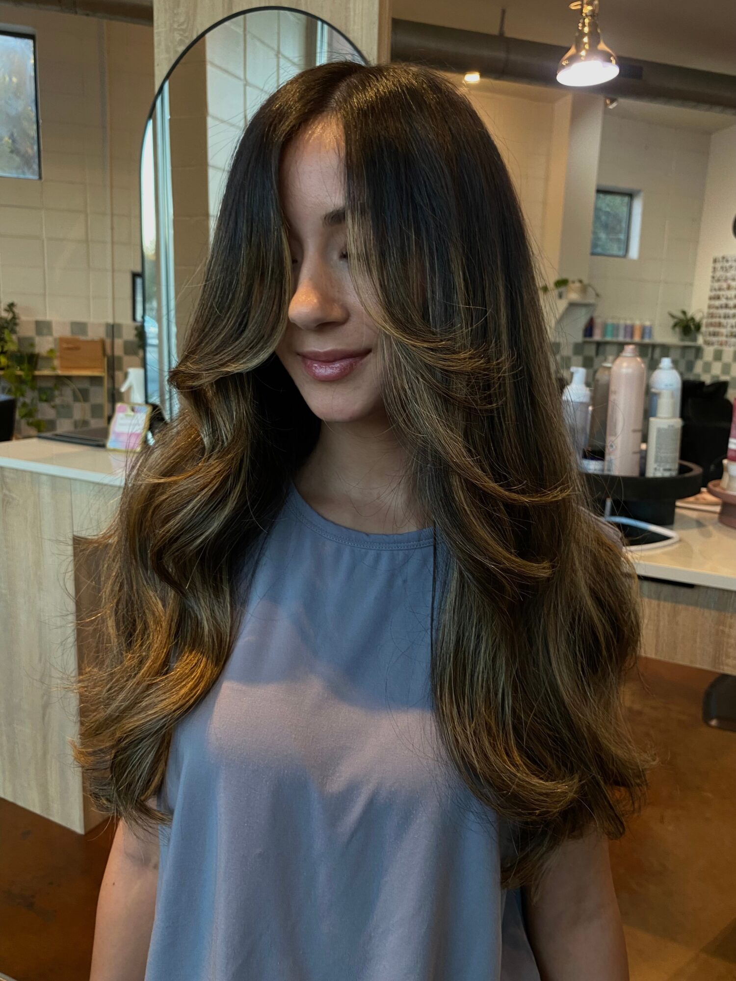 Person with long, wavy brown hair seated in a salon, wearing a blue-gray cape.