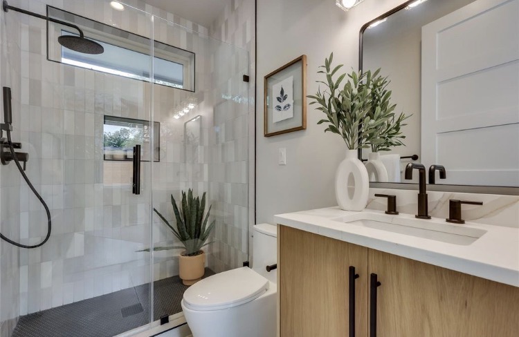 A tiled bathroom with a glass shower, toilet, wooden vanity, sink, mirror, and potted plants.