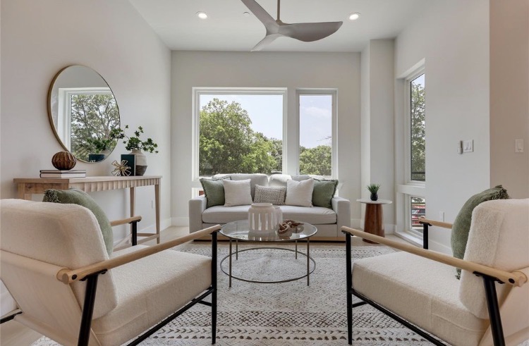 Bright living room with two cream chairs, a glass coffee table, large windows, plants, and a ceiling fan.